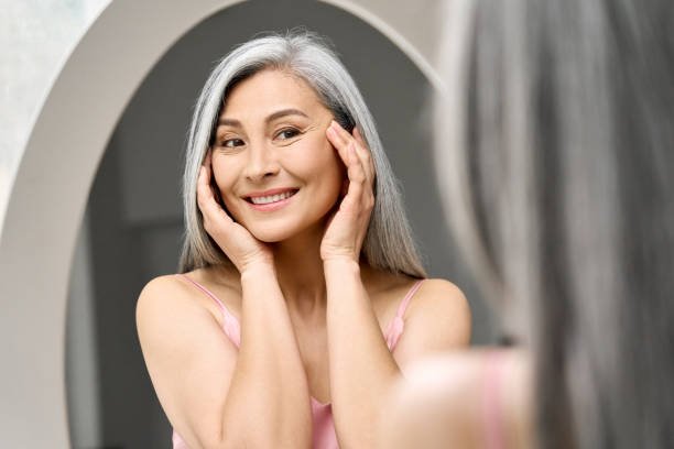 Happy middle 50 years aged asian woman with gray hair looking at mirror reflection examining touching face enjoying antiaging beauty treatments. Beauty hydrate skin care wrinkle prevention concept.
